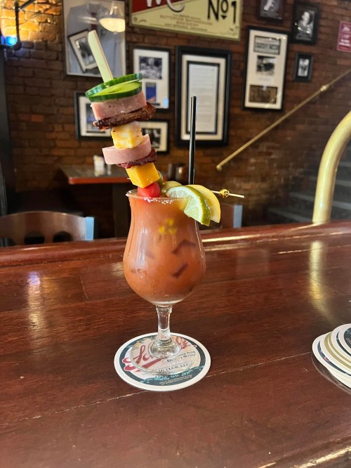 A colorful cocktail garnished with a skewer of meats and vegetables, served in a glass on a wooden bar.