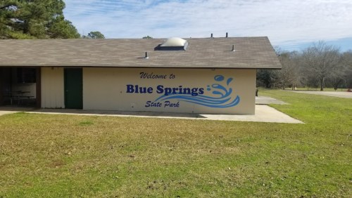 A building with a sign that reads "Welcome to Blue Springs State Park," surrounded by grass and trees.