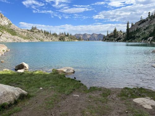 A serene lake surrounded by mountains and trees, with clear blue water and a partly cloudy sky.