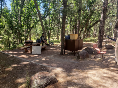 A campsite in a wooded area with a picnic table, chairs, and a person sitting nearby among trees and rocks.