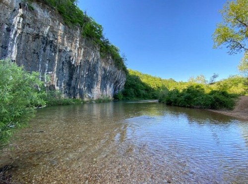 A serene river flows beside a rocky cliff, surrounded by lush greenery under a clear blue sky.