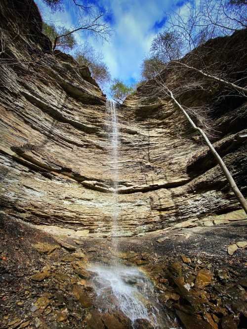 A waterfall cascades down a rocky cliff surrounded by trees under a blue sky with scattered clouds.