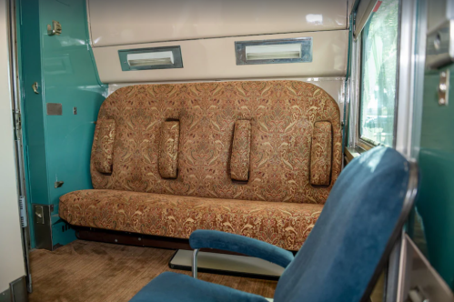 Interior of a vintage train car featuring a patterned sofa and a blue armchair against a teal wall.