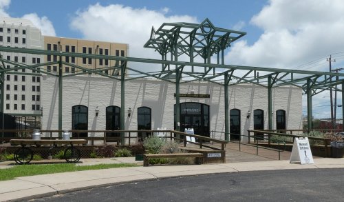 A modern building with a metal framework, large windows, and a wooden deck, set against a blue sky with clouds.