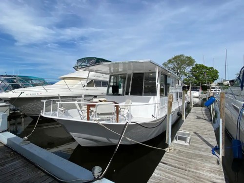 A white houseboat docked at a marina, with a wooden deck and chairs, surrounded by other boats and a clear blue sky.