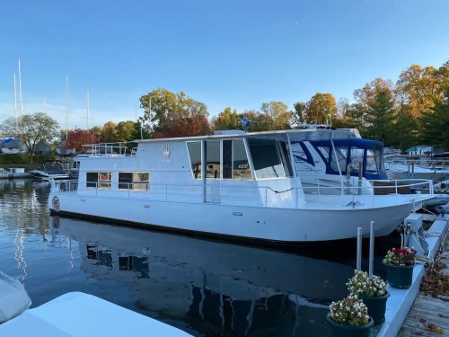 A white houseboat docked at a marina, surrounded by trees and other boats under a clear blue sky.