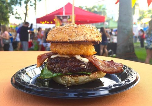 A close-up of a sesame seed burger with bacon, lettuce, and a crispy onion ring, served on a black plate.
