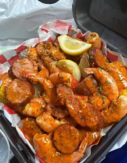 A takeout container filled with shrimp, sausage, corn, and lemon wedges, all coated in a spicy sauce.