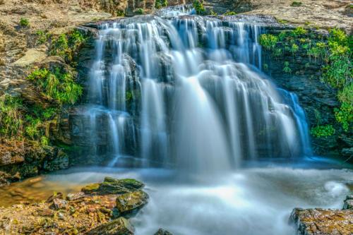 A serene waterfall cascades over rocks, surrounded by lush greenery and a tranquil pool below.