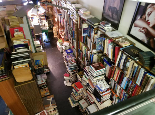 A cozy room filled with tall bookshelves overflowing with books, some stacked on the floor, and a ceiling fan above.