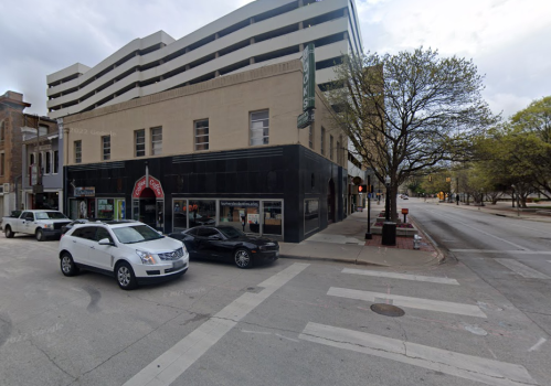 Street view of a city intersection with parked cars, a building with a sign, and trees lining the sidewalk.