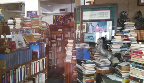 A cluttered bookstore filled with stacks of books, shelves, and various decorative items, creating a cozy, chaotic atmosphere.