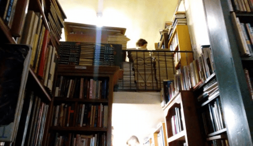 A cozy bookstore interior with shelves of books and a person on a balcony above, illuminated by soft light.