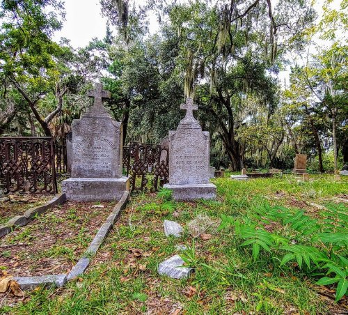 Two weathered gravestones in a grassy cemetery, surrounded by trees and moss, with a peaceful, serene atmosphere.