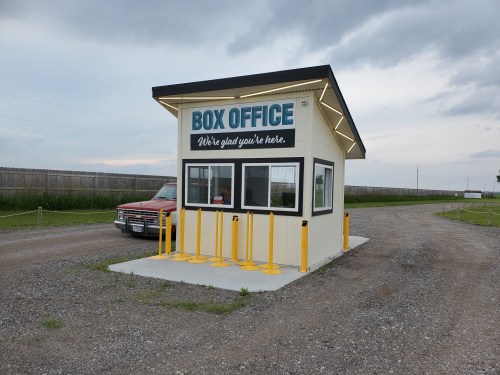 A small box office building with a sign reading "Box Office" and a red vehicle parked nearby on a gravel road.