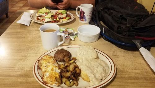 A breakfast plate with eggs, sausage, potatoes, and gravy, alongside a coffee cup and a plate of salad in the background.