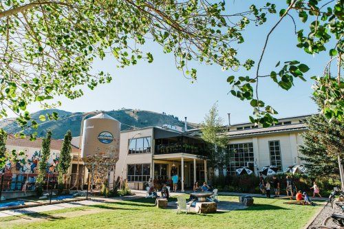 A vibrant outdoor space with people enjoying a sunny day near a brewery and scenic mountains in the background.