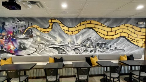 A vibrant mural featuring a cup, a bridge, and a city skyline, with yellow brick patterns and artistic swirls.