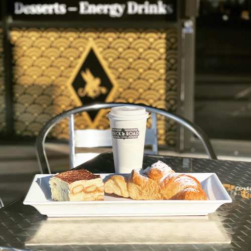 A plate with a croissant and dessert, alongside a coffee cup, on a table outside a café.