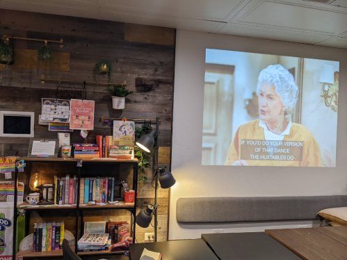 A cozy room with a projector displaying a scene from a show, surrounded by books and decor on the wall.