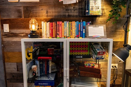 A bookshelf filled with colorful books and board games, illuminated by a vintage lamp, against a wooden wall.