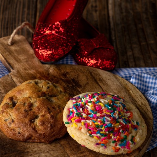 A wooden board with two cookies: one chocolate chip and one with colorful sprinkles, alongside red glittery shoes.