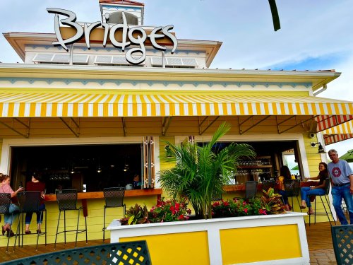 A colorful restaurant named "Bridges" with a striped awning, outdoor seating, and tropical plants in the foreground.