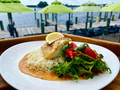 A plate of fish with lemon, served on rice and a fresh salad, with a waterfront view and green umbrellas in the background.