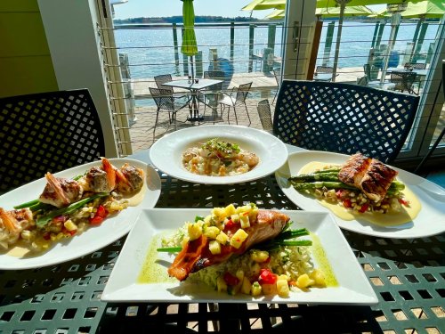 A table with four beautifully plated seafood dishes, featuring fish, vegetables, and a scenic waterfront view.