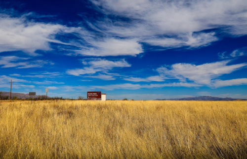 A vast field of golden grass under a blue sky with clouds, featuring a distant sign and buildings.