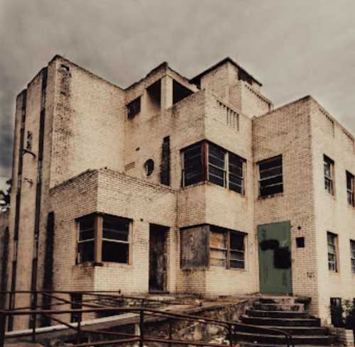 An abandoned, weathered brick building with broken windows and a green door, surrounded by overgrown vegetation.