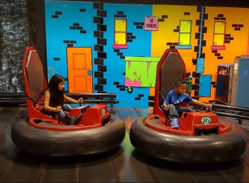 Two children in red bumper cars, set against a colorful, graffiti-style background with a "No Parking" sign.