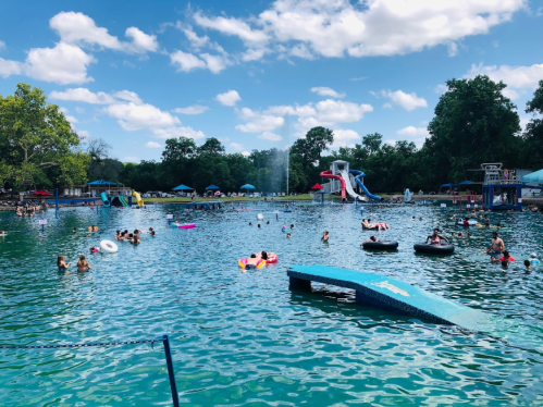 A lively swimming area with people enjoying the water, colorful floats, and a water slide under a blue sky.