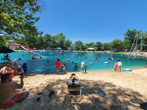 A sunny day at a swimming area with people enjoying the water, sandy beach, and trees in the background.