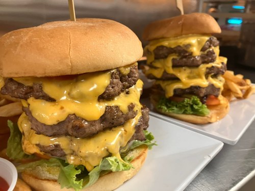 A towering cheeseburger with multiple beef patties, melted cheese, lettuce, and tomato, served with fries.