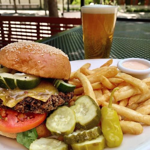A delicious burger with cheese, pickles, and tomato, served with fries and a cold beer on a table outdoors.