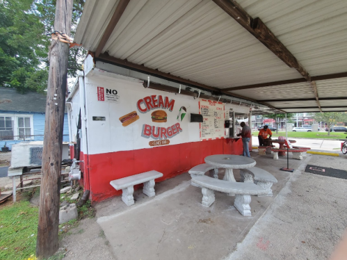 A small outdoor eatery with a red and white facade, featuring a sign that reads "Cream Burger" and stone picnic tables.