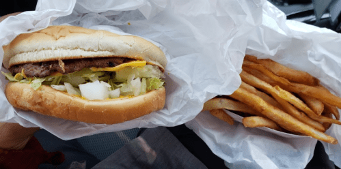 A hand holding a hamburger with lettuce, onions, and cheese, alongside a container of golden French fries.