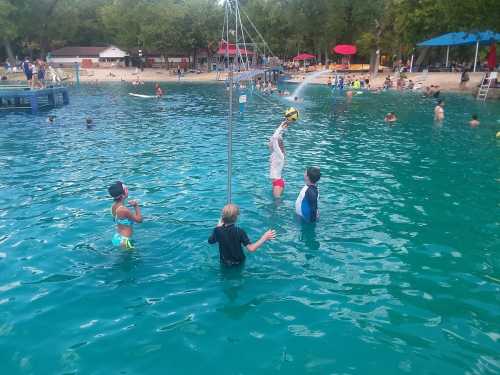 Children playing in a turquoise pool, with one jumping to catch a ball and others enjoying the water.