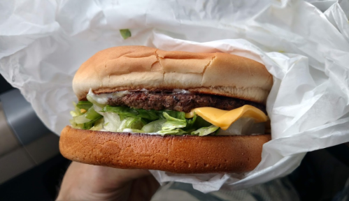A hand holding a cheeseburger with lettuce, cheese, and a soft bun, wrapped in white paper.