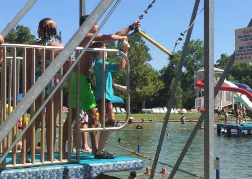 Children on a platform at a water park, preparing to use a zip line over a swimming area with slides in the background.