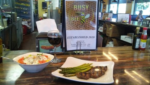 A plate with grilled meat, asparagus, and a salad, alongside a glass of wine, with a "Busy Bee" sign in the background.