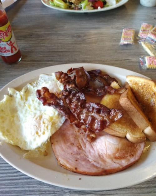 A hearty breakfast plate with eggs, bacon, ham, toast, and hot sauce on the side.