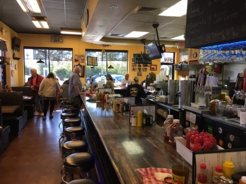A cozy diner interior with a long counter, stools, and patrons enjoying their meals. Bright yellow walls and condiments visible.