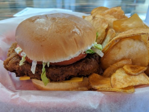 A crispy chicken sandwich with lettuce, tomato, and onion, served with a side of potato chips in a basket.