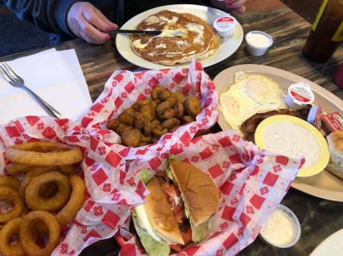 A table with a plate of pancakes, a breakfast platter, onion rings, and a sandwich, all served with dipping sauces.
