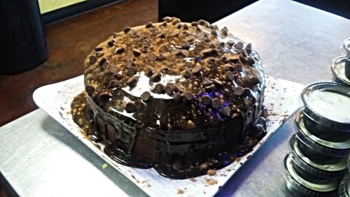 A rich chocolate cake topped with chocolate chips and drizzled with chocolate sauce, served on a white plate.