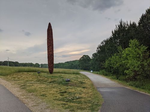 A tall, slender sculpture stands in a grassy area beside a winding path, with trees and a cloudy sky in the background.