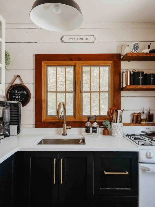 A cozy kitchen with a white countertop, black cabinets, wooden shelves, and a large window letting in natural light.