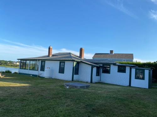 A white house with green trim and a large yard, set against a clear blue sky and near a body of water.